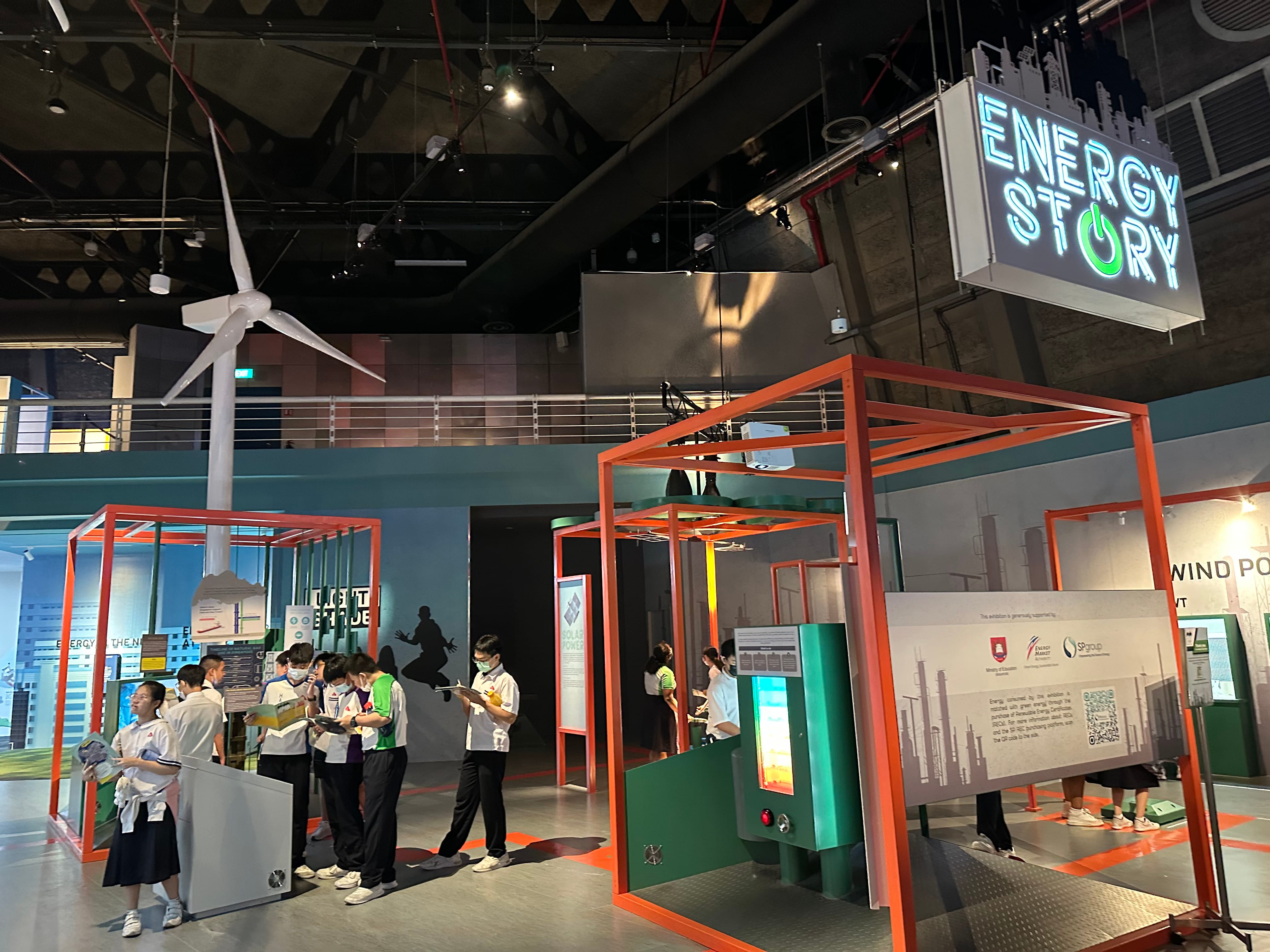 Students explore the Energy Story museum exhibit with orange frames, interactive displays, and a wind turbine model overhead.